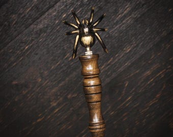 May include: A bottle stopper featuring a dark metallic spider topper with gold-toned highlights. The handle is crafted from polished brown wood with a series of horizontal grooves, leading down to a tapered silver-colored metal base component.