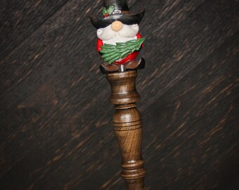 May include: A bottle stopper featuring a gnome topper with a black hat, white beard, red clothing, and green pine branches. The gnome sits atop a turned dark brown wooden handle with a smooth, polished finish and a silver-toned metal base.