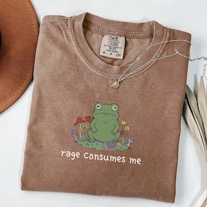 Puede incluir: Camiseta Comfort Colors marrón con una ilustración de una rana verde rodeada de flores y setas. El texto "rage consumes me" está impreso debajo. Un collar plateado con un colgante está sobre la camiseta.