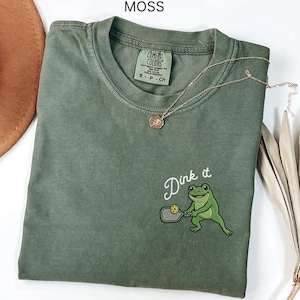 Puede incluir: Una camiseta verde musgo con el texto "Dink it" y un bordado de una rana jugando al pickleball. La camiseta tiene una etiqueta Comfort Colors y un collar dorado con un colgante. La camiseta está doblada sobre una superficie blanca.