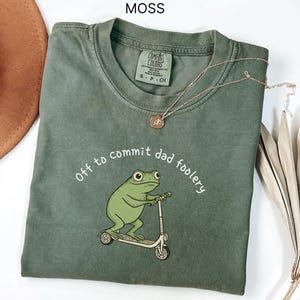 May include: A moss green t-shirt with the text "Off to commit dad foolery" and a cartoon frog riding a scooter. The shirt has a crew neck and short sleeves. A gold necklace with a pendant is draped across the shirt.