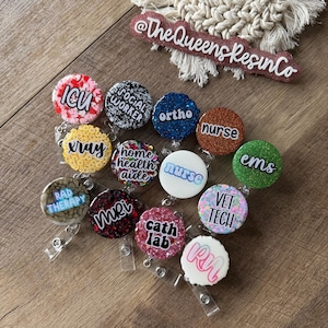 May include: A collection of round, colorful badge reels with various job titles like "ICU," "Nurse," and "Vet Tech." Each reel features glitter and text, with a clear plastic clip. The background includes a wooden surface and a decorative sign that reads "@TheQueensResinCo."