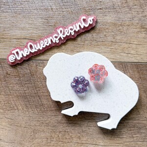 May include: A white buffalo-shaped display board with two paw-shaped resin charms. One charm is purple with glitter, and the other is pink with glitter and the text LARK. A pink glittery sign with white text reads @TheQueensResinCo.