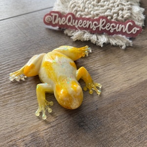 May include: A resin frog figurine in shades of yellow and white with glitter accents. The frog is lying down with its legs outstretched. The text "@TheQueensResinCo" is visible in the background.