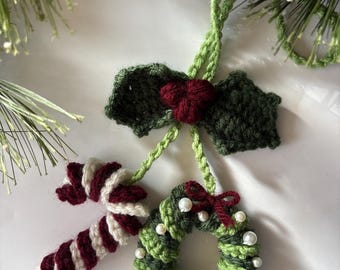 May include: Handmade Christmas decorations. The image shows a crocheted candy cane, a holly leaf with a red berry cluster, and a wreath decorated with white beads and a red bow. The colours are green, white, and burgundy.