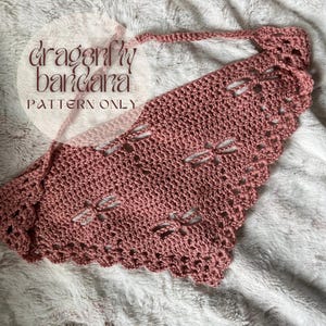 May include: A pink crocheted bandana with a dragonfly design. The bandana has a scalloped edge and is tied with a long string.