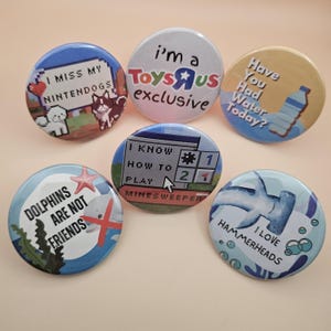 Op de afbeelding: Zes ronde buttons met verschillende ontwerpen en tekst. Eén button zegt "I MISS MY NINTENDOGS" met gepixeleerde hondengraphics. Een andere zegt "I'm a Toys R Us exclusive". Andere buttons hebben "Dolphins are not friends" en "I love Hammerheads".