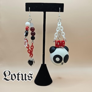 Puede incluir: Un par de pendientes desiguales. Uno presenta una cabeza de panda con la palabra "love" encima, y el otro tiene un diseño de cadena y cuentas con un dije de corazón. Los pendientes se exhiben en un soporte negro.