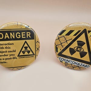 May include: Two circular badges with a yellow and black design. One features the word "DANGER" and a radiation symbol, with the text "Interaction with this object could render you to a post-life state. COGNITOHAZARD." The other says "COGNITOHAZARD."