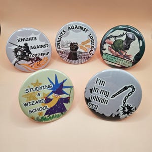 Puede incluir: Cinco botones redondos con varios diseños y texto. Uno presenta un caballero a caballo con las palabras "Knights Against Lordship". Otro muestra un puño y trigo con el mismo texto. Otros botones tienen un mago, una rana y la frase "I'm in my villain era."