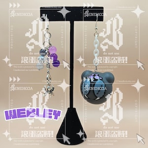 May include: A pair of mismatched earrings hanging on a black T-shaped stand. The left earring features a silver-toned barbed wire charm, a small spider charm, and purple and grey beads. The right earring features a white plastic chain and a round grey bear-shaped charm with a spider-like face design.