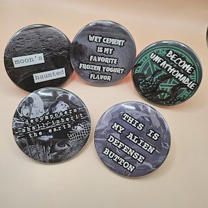 May include: A collection of five round buttons with various designs and text. One button features a moon and the words "moon's haunted." Another says "Wet Cement is my favorite frozen yogurt flavor." Other buttons have the phrases "Become Unfathomable," "decomposers shall inherit the earth," and "This is my alien defense button."