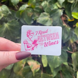 Op de afbeelding: Een witte sticker met roze tekst die "I Read Between the Wines" zegt. Een cartoonvrouw in een roze jurk houdt een glas wijn en een boek vast.