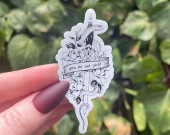 May include: Black and white sticker with a dagger, snake, and flowers. The sticker has the text "you do not yield" on a banner.