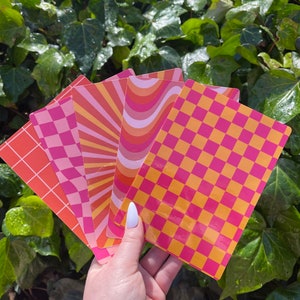 May include: Five colorful patterned sheets of paper with pink, orange, and yellow geometric designs. The sheets are arranged in a fan shape and held in a hand.