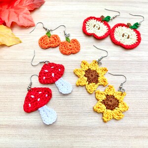 May include: Crocheted earrings in various autumnal themes, including pumpkins, apples, mushrooms, and sunflowers. The earrings are displayed on a wooden surface.