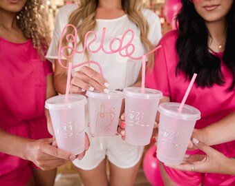 May include: Four friends holding clear plastic cups with pink straws and pink glitter writing that says "Bride Tribe". One straw is shaped into the word "Bride".