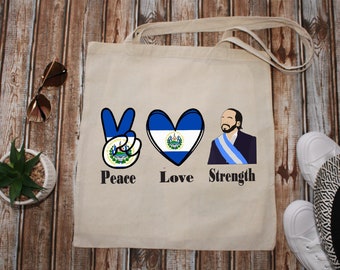 May include: Beige tote bag featuring a peace sign, a heart, and a portrait, all incorporating the flag of El Salvador. The words "Peace," "Love," and "Strength" are printed below the images. Sunglasses, a small plant, and shoes are visible.