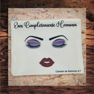 May include: A white canvas makeup bag with a zipper closure. The bag features a stylized face with closed purple eyeshadow, long eyelashes, and red lips. The text "Eres Completamente Hermosa" and "Cancion de Solomon 4:7" are printed on the bag.