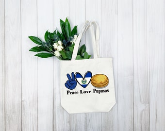 May include: A white canvas tote bag with the words "Peace Love Pupusas" printed on it. The design includes a peace sign, a heart with the El Salvador flag, and a stack of pupusas. Green foliage and white flowers are visible.