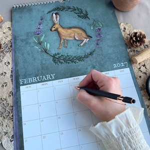 Könnte beinhalten: Ein Wandkalender für 2027, geöffnet für Februar. Der Kalender zeigt eine Aquarellillustration eines braunen Hasen, umgeben von einem floralen Kranz. Das Wort „FEBRUAR“ steht oben, darunter die Wochentage. Eine Hand hält einen Stift.