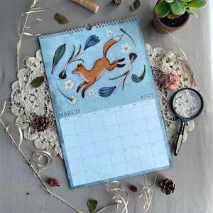 May include: A March 2027 calendar with a leaping fox illustration and floral details. The calendar is spiral-bound and set against a light blue backdrop. Decorative elements include a magnifying glass, a small plant, and other natural items.