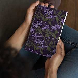 Puede incluir: Cuaderno de espiral con una cubierta morada oscura que presenta un patrón repetido de colibríes morados, flores blancas y hojas verdes. El cuaderno está abierto, revelando páginas blancas.