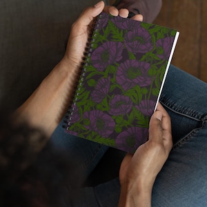 May include: A spiral-bound notebook featuring a floral pattern. The design showcases large purple flowers and green leaves against a dark background. The notebook is held open, revealing blank white pages.