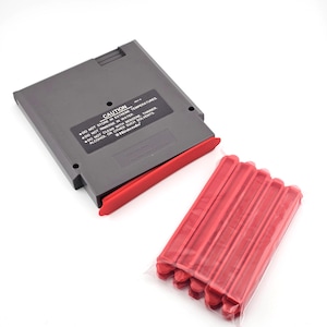 May include: A gray rectangular video game cartridge with a black label featuring white text that reads CAUTION, DO NOT STORE IN EXTREME TEMPERATURES, DO NOT IMMERSE IN WATER, DO NOT CLEAN WITH BENZENE, THINNER, ALCOHOL OR OTHER SUCH SOLVENTS, Nintendo. A red plastic clip is attached to the cartridge, and a set of five additional red plastic clips is enclosed in a clear plastic bag.