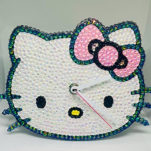 May include: A white Hello Kitty clock with a pink bow and black eyes. The clock is made of rhinestones and has a green border.