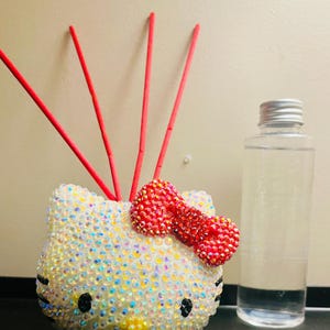 May include: A white Hello Kitty diffuser with a red bow and clear rhinestones. The diffuser has four red sticks and is next to a clear glass bottle.