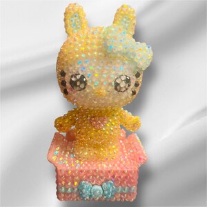 May include: A yellow and pink sparkly rabbit figurine sitting in a pink sparkly box with a blue bow. The rabbit has black eyes and a blue bow on its head.
