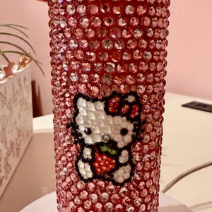 Puede incluir: Un vaso rojo cubierto de brillantes pedrería. El vaso presenta un diseño de Hello Kitty sosteniendo una fresa. La tapa es roja y tiene un cierre abatible. El vaso está diseñado para contener bebidas frías.