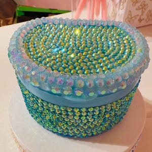 May include: A small, oval-shaped container covered in iridescent rhinestones. The container is primarily turquoise, with a light blue rim and band. The rhinestones reflect various colors, creating a sparkling effect. The container sits on a white surface.