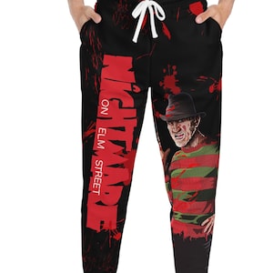 Op de afbeelding: Zwarte joggingbroek met een rood en zwart grafisch ontwerp. De tekst "NIGHTMARE ON ELM STREET" staat verticaal op de linkerpijp. De rechterpijp heeft een afbeelding van Freddy Krueger.