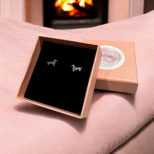 Könnte beinhalten: Ein Paar silberne Dackel-Ohrringe in einer kleinen braunen Schachtel mit schwarzem Innenraum. Die Schachtel hat ein weißes Etikett mit dem Text "Handmade by Riddle".