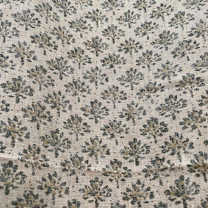 May include: Close-up of a beige fabric with a repeating pattern of stylized, dark green floral motifs. The fabric has a slightly textured appearance, suggesting a natural fiber like linen or cotton. The floral design is evenly spaced across the surface.