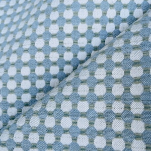 May include: Close-up of a light blue fabric with a repeating pattern of white and light blue circles. The fabric has a textured appearance, with a subtle grid-like structure visible between the circles. The material is folded, showing the fabric's thickness.