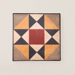 May include: A square tile with a geometric pattern in shades of brown, black, orange, and red. The tile is framed with a black border.