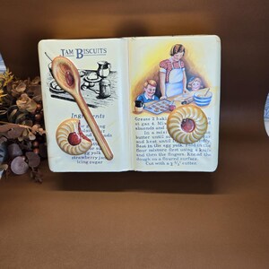 May include: A vintage cookbook with a yellow cover and a recipe for jam biscuits. The cookbook is open to the recipe page, which includes a list of ingredients and instructions. The image also shows a wooden spoon and a plate of jam biscuits.