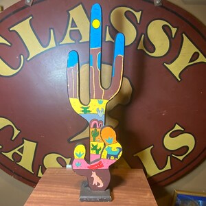 May include: A colorful wooden cactus sculpture with a blue sky, yellow sun, and desert landscape painted on it. The cactus is standing on a wooden base.
