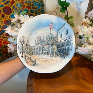 May include: White ceramic plate with a detailed illustration of a European city scene, featuring buildings, streetlights, and people. The plate has the words "Post Card" printed on it. The plate is held in front of a decorative background.
