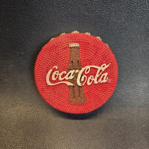 May include: A red round button with a white Coca-Cola logo and a brown bottle graphic.