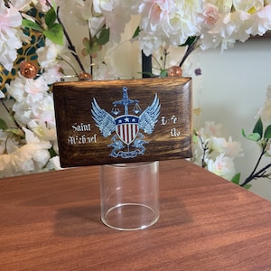 May include: A wooden box with a Saint Michael design. The box has a dark brown stain and features a winged shield with a sword, scales of justice, and an American flag. The text "Saint Michael" is written on the left side of the box and "L.F.U." is written on the right side.