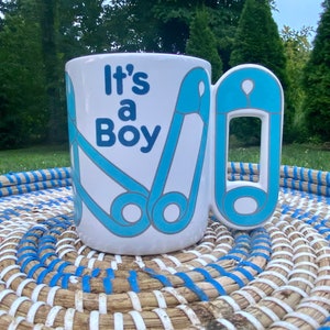 May include: White ceramic mug with a blue and white safety pin design. The mug has the text "It's a Boy" on the front.