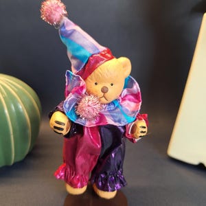 May include: A small, brown teddy bear dressed as a clown. The bear is wearing a multi-colored hat, a pink, purple, and blue ruffled collar, and a pink and purple jester costume. The bear is standing on a brown metal base.