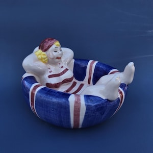 May include: A ceramic figurine of a person reclining in a blue and white striped inner tube. The person is wearing a red and white striped swimsuit and a red cap. The inner tube has a blue background with white and red stripes.