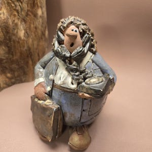 May include: A ceramic figurine of a person holding a phone and a briefcase. The figurine is painted in shades of blue, brown, and black. The person has a surprised expression on their face.