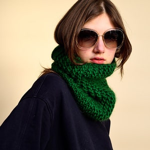 Könnte beinhalten: Eine Frau trägt einen dunkelblauen Pullover mit einem grünen gestrickten Halstuch und einer großen, runden, braunen Sonnenbrille.