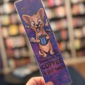 May include: A rectangular bookmark featuring a cartoon dog holding a blue coffee mug, surrounded by floating books against a purple and blue cosmic background. The text reads, "powered by COFFEE & plot twists" in dark purple and black lettering.